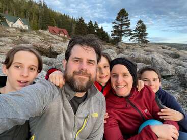 Das Bild zeigt eine vierköpfige Familie, die für ein Selfie an einem felsigen Strand posiert, mit einem Haus und Bäumen im Hintergrund unter einem bewölkten Himmel.