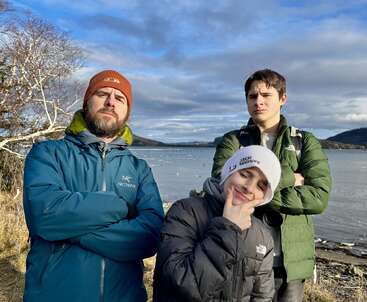 Trois personnes posant avec des expressions enjouées et confiantes au bord d'un lac pittoresque. Elles portent des vestes et des chapeaux d'hiver. La lumière du jour, le ciel nuageux et les arbres dénudés complètent le paysage.