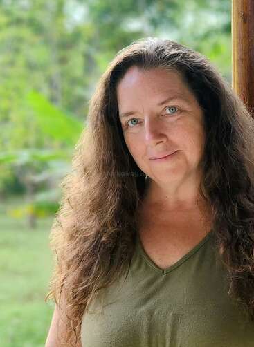 A woman with long, wavy brown hair and blue eyes smiles gently. She's wearing a green top and standing outdoors with lush greenery in the background.