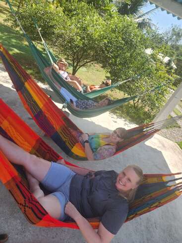 Four people relax in colorful hammocks outdoors, smiling and enjoying the sunny day. Lush green trees and a bright sky create a peaceful, cheerful atmosphere.
