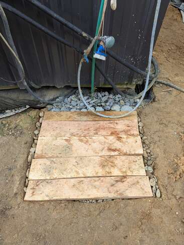 Una ducha improvisada al aire libre con tablones de madera sobre rocas, mangueras, una alcachofa de ducha, un cepillo y una pared negra con tuberías a la vista y suelo de tierra.