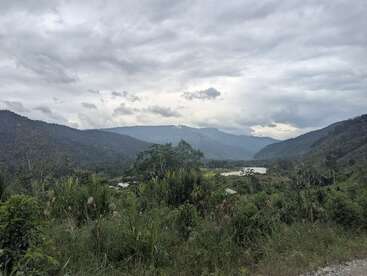 Un exuberante valle verde se extiende entre dos cadenas montañosas bajo un cielo nublado, con una densa vegetación en primer plano y un pequeño río serpenteando.