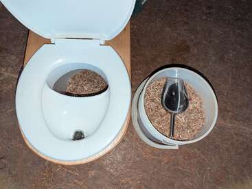 Esta imagen muestra un inodoro de compostaje con virutas de madera dentro de la taza. Al lado, hay un cubo con virutas adicionales y una pala para mayor comodidad.