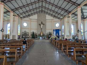 El interior de una iglesia espaciosa con bancos de madera, vidrieras, un gran crucifijo y adornos navideños. Varias personas están sentadas, rezando o reflexionando en silencio.
