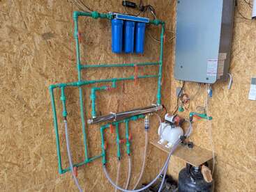 Esta imagen muestra un sistema de filtración de agua con cartuchos de filtro azules, tuberías de PVC verdes, válvulas, una bomba y un panel de control montado en una pared de madera.