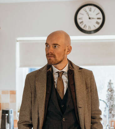 La imagen muestra a un hombre trajeado de pie en una cocina, con un reloj en la pared detrás de él y una ventana a su derecha.