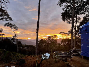 A vibrant sunset illuminates the sky behind tall trees in a forest. Foreground shows dirt, scattered objects, a mirror reflecting sky, and a blue tent.