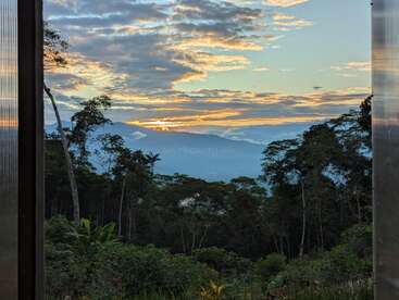 Golden sunlight illuminates misty mountains and lush rainforest, framed by two doorways. Dramatic clouds float above, creating a tranquil, breathtaking natural scene at dawn or dusk.