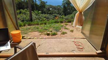 An open doorway reveals a lush garden and forest view. Inside, there’s a chair, a laptop, orange bucket, curtain, and an extension cord. Bright sunlight streams in.