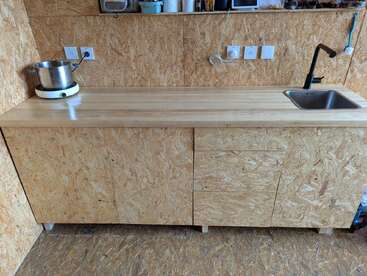 This image shows a simple kitchen counter made of oriented strand board (OSB) with a stainless steel sink, electric hot plate, and minimal, rustic decor.