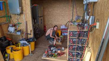 A person crouches and smiles in a workshop filled with tools, batteries, wires, yellow buckets, and various equipment, appearing to prepare food or work on a project.
