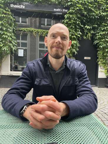 A man sits at an outdoor table with hands clasped, smiling. Behind him, green ivy covers a building with a "sushi x thai" restaurant sign.