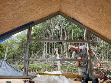 A person is building or installing a window frame in a triangular wooden structure. Outside, dense green forest is visible. Construction tools and materials are nearby.