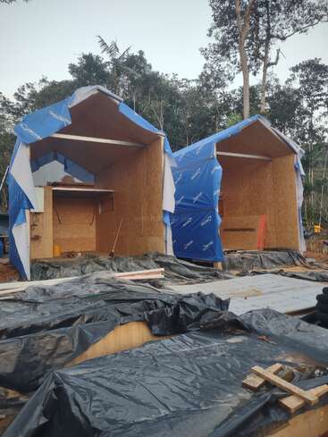 Zwei kleine, im Bau befindliche Holzkonstruktionen sind mit blauen Schutzplanen abgedeckt. Schwarze Plastikplanen und verschiedene Baumaterialien sind auf dem Boden verteilt, umgeben von Bäumen.