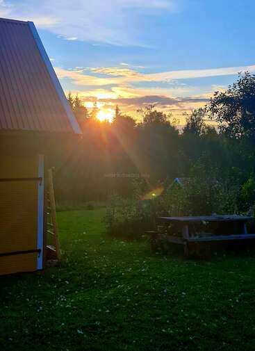 The sun sets behind tall trees, casting golden rays across a green lawn. A wooden house, ladder, and picnic table create a peaceful countryside atmosphere.