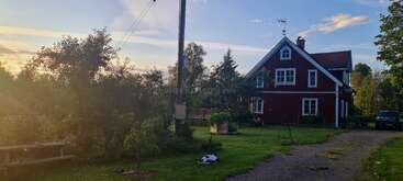 A peaceful countryside scene features a red house with white trim, surrounded by greenery, trees, a parked car, garden furniture, and soft sunlight casting gentle shadows.