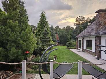 Uma cena tranquila no quintal, com árvores verdejantes, um gramado bem cuidado, cadeiras reclináveis modernas, uma casa branca com detalhes em pedra e um céu nublado ao pôr do sol.