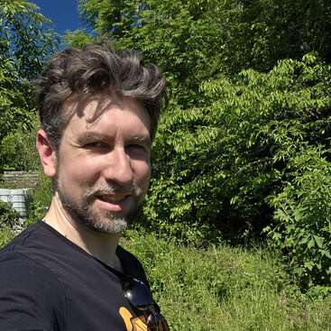 Um homem de cabelo castanho e barba está sorrindo ao ar livre, vestindo uma camisa preta. Ele está ao lado de um cortador de grama, cercado por uma vegetação verdejante e pela luz do sol.