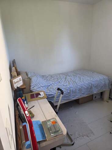 Um quarto pequeno e arrumado tem uma cama bem feita com lençóis listrados de azul, uma mesa de madeira com livros e artigos de papelaria, uma cadeira e um tapete de cor clara.