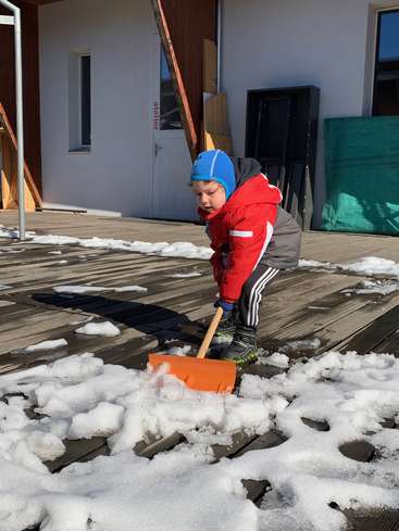 A young child in winter attire is shoveling snow on a wooden deck outside a building with a white door and a window.