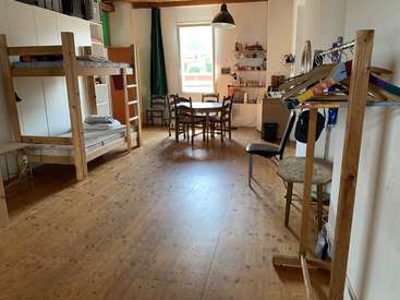 The image depicts a room with bunk beds, a dining table, and a desk, featuring wooden floors and walls, with a window and various furniture and objects scattered throughout.