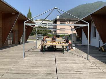 The image depicts a group of people working on a metal frame in an outdoor setting, surrounded by buildings and trees.