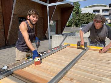 Two men are working on a wooden structure, one holding a drill and the other a level, in a yard with trees and buildings in the background on a sunny day.