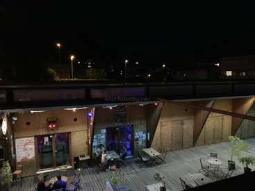 The image depicts a serene outdoor seating area at night, featuring tables and chairs on a wooden deck, with a building in the background and a dark sky above.