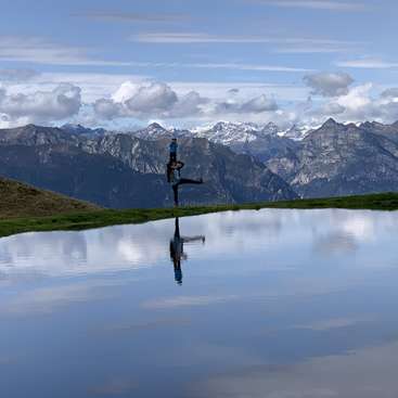 The image depicts a serene landscape featuring a person in a yoga pose on the edge of a tranquil lake, set against a majestic mountain range under a blue sky with clouds.