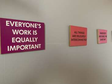 The image features three pink signs with white and black text on a white wall, conveying messages of equality and interconnectedness. The signs are rectangular and appear to be made of wood.