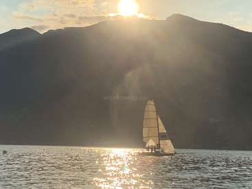 A sailboat glides across the water, its white sails billowing in the wind, set against a majestic mountain range and a radiant sun shining brightly in the sky.