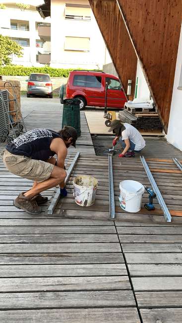 Two people are working on a wooden deck, surrounded by tools and materials, with a red van and building visible in the background on a sunny day.