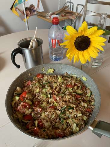The image depicts a table with a pan of food, a sunflower, and a water bottle, set against a white wall with a chair and other objects in the background.