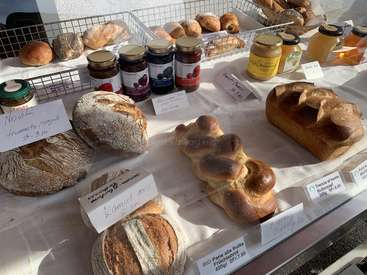 The image shows a table with various types of bread and jars of food on display, likely at a bakery or market. The items are arranged neatly and have price tags.