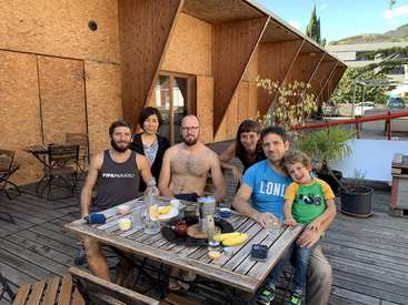 The image depicts a group of six people, including two children, gathered around a wooden table on a deck, with a building featuring wood paneling in the background.