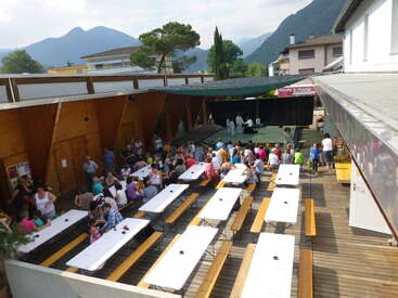 An outdoor gathering with people seated at long tables, some watching a stage event. Surrounded by mountains and buildings, the atmosphere is lively and communal.
