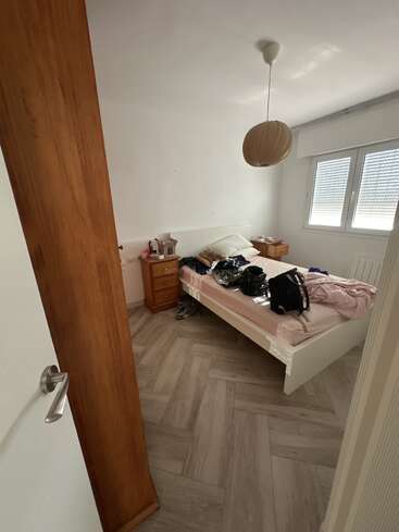 Um quarto pequeno com uma cama branca, um pouco de bagunça em cima, duas mesinhas de cabeceira de madeira, uma janela que deixa entrar luz e uma lâmpada moderna pendurada. A porta está entreaberta.