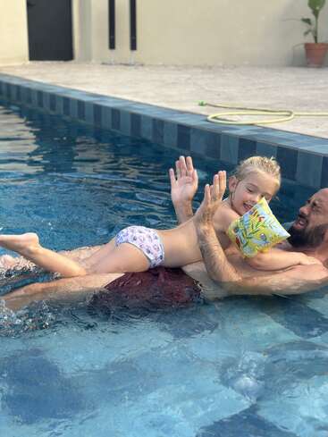 Um homem e uma criança estão brincando alegremente em uma piscina. A criança usa bóias e fralda de natação, e ambos parecem estar rindo e se divertindo.