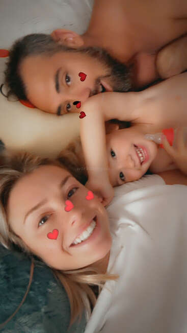 Una familia feliz de tres miembros se acurruca sonriente y relajada. Filtros de corazones rojos decoran sus caras. El niño sostiene un chupete, creando un ambiente cálido y cariñoso.