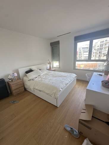 Il s'agit d'une chambre lumineuse et minimaliste avec du parquet, un lit blanc, une table de chevet, une commode, des pantoufles, des boîtes et de grandes fenêtres donnant sur les bâtiments modernes de la ville.