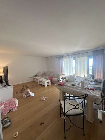 Un salon confortable et lumineux avec du parquet. Un enfant joue par terre parmi ses jouets. On aperçoit une table à manger en désordre et un canapé. Les rideaux diffusent la lumière du jour.