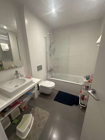 Cette image montre une salle de bain moderne avec un lavabo, des toilettes, une baignoire-douche, un chariot de rangement, un carrelage gris, un meuble sous lavabo, un tapis et un éclairage lumineux.