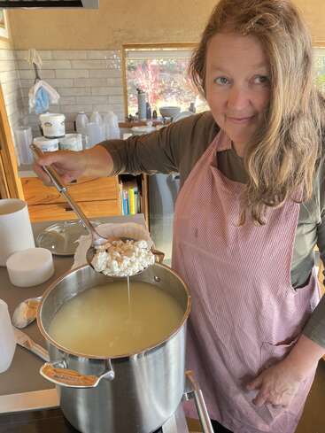 A woman in a striped apron is making cheese in a kitchen, holding a ladle of curds over a large pot filled with whey. She’s smiling.