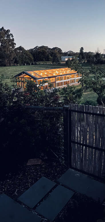 A glowing greenhouse sits in an open green field, surrounded by trees and bushes, viewed through a rustic wooden gate at dusk with a peaceful atmosphere.