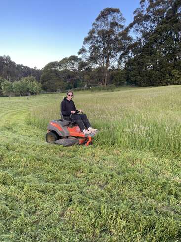 A person wearing sunglasses and dark clothing is riding a sit-on lawnmower, cutting tall grass in a large, green field surrounded by trees.