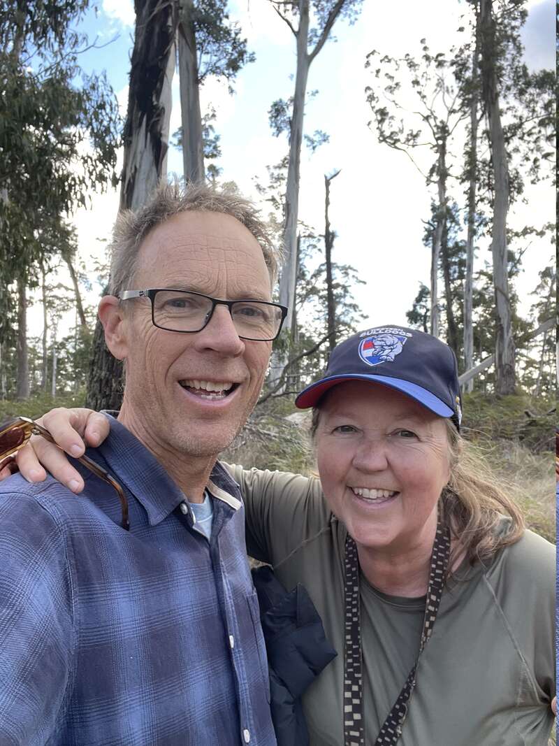 A smiling man and woman take a selfie outdoors in a forest. The man wears glasses, the woman a cap. Both appear happy, enjoying nature together.