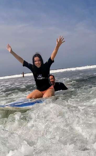 Une jeune fille rayonnante est agenouillée sur une planche de surf dans les vagues peu profondes de l'océan, les bras levés dans un élan d'enthousiasme, tandis qu'un homme la soutient par derrière. Une scène de plage lumineuse et ensoleillée.