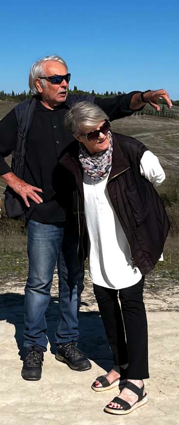 An older man and woman stand outside on a sunny day. He gestures toward the distance while she looks, both wearing sunglasses and casual clothing.