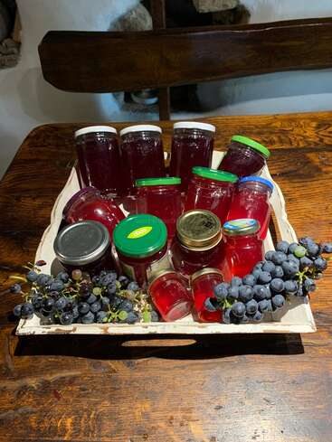 A wooden tray holds various jars of homemade grape jelly, some with colorful lids. Fresh grape bunches are placed on both sides, all on a rustic table.