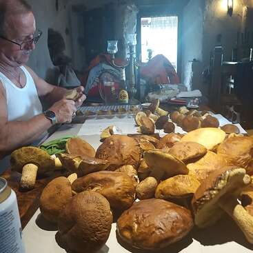 A man in a white tank top cleans mushrooms at a rustic table filled with large, fresh mushrooms in a cozy, warmly lit kitchen setting.